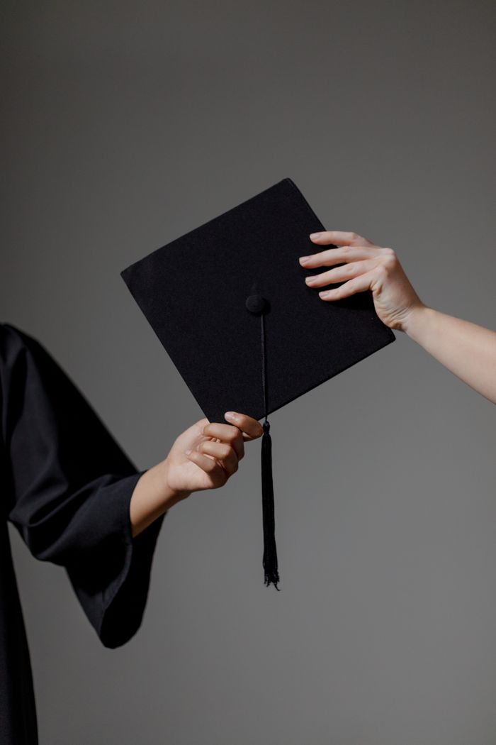 Two people exchanging a graduation cap on a grey background, symbolizing achievement.